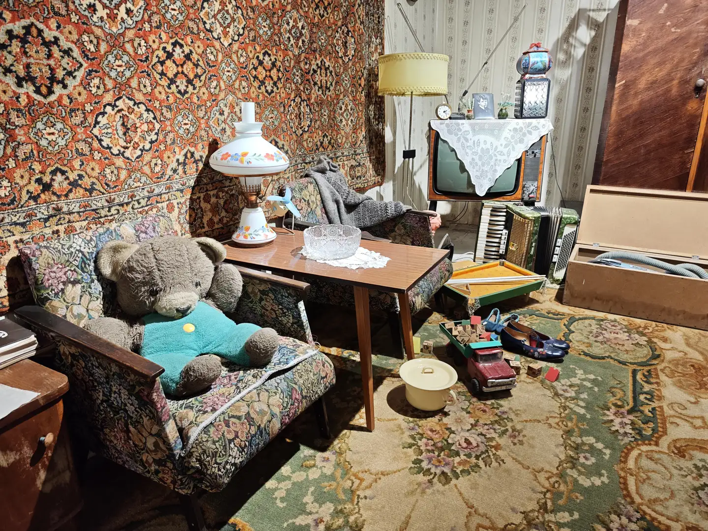 客厅里摆放着花纹沙发和木质茶几，茶几上有一盏装饰灯和玻璃碗，墙上挂着色彩丰富的挂毯，地上散落着玩具车、积木和小锅。电视柜上覆盖着蕾丝桌布，旁边放着一台老式电视机和手风琴，角落里还有一个打开的木箱。