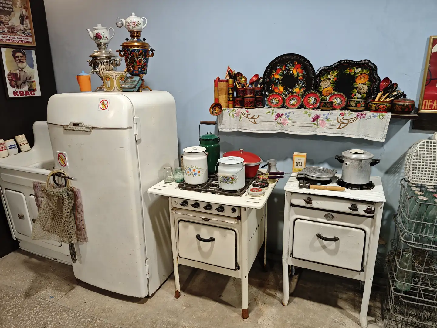 厨房里陈列着一台白色老式冰箱，上面放置着茶壶和金属容器，旁边是两个小型炉灶，上面有锅具和搪瓷罐，墙上的架子摆满了彩绘盘子和厨具。