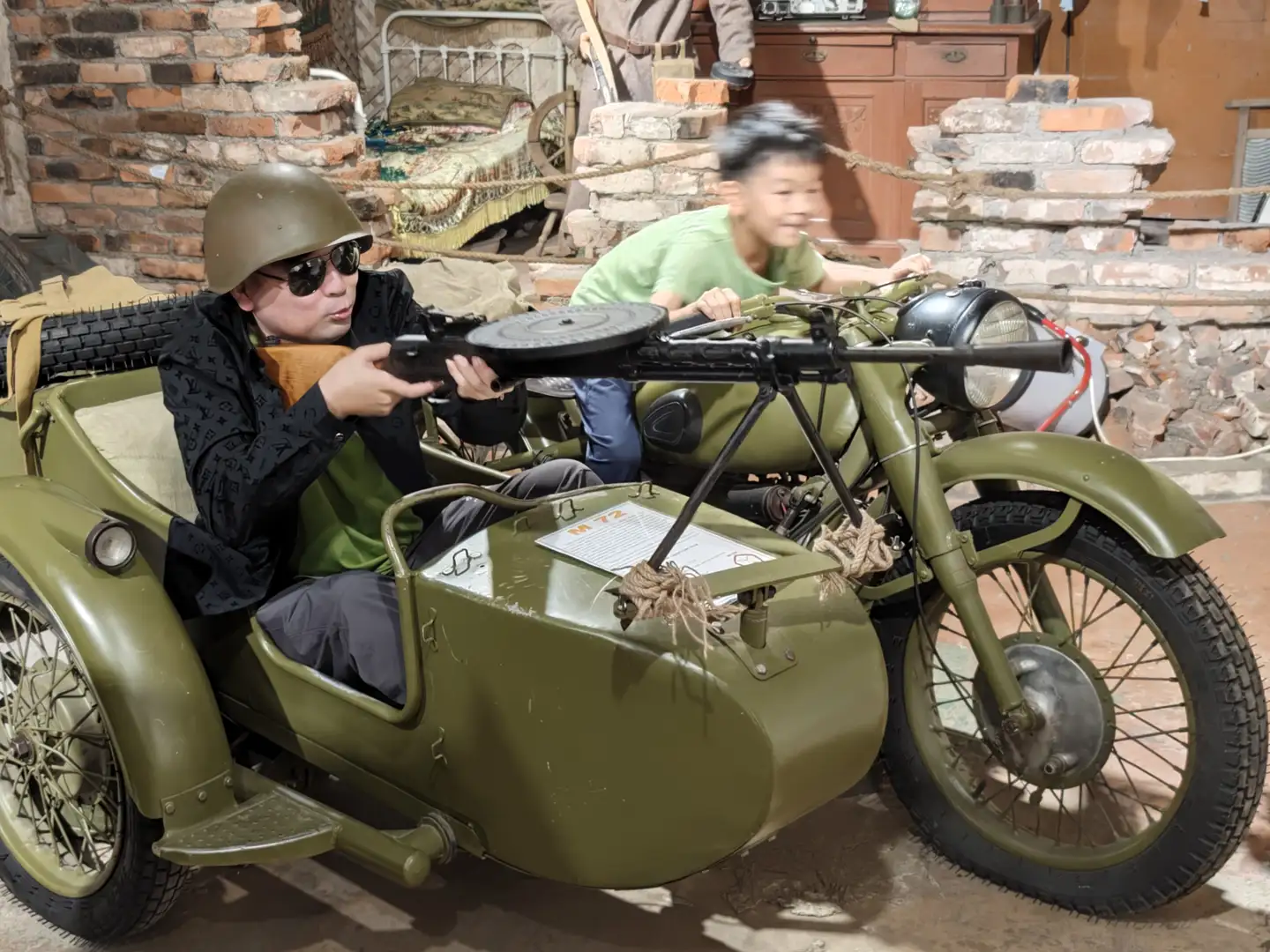 一辆绿色军用摩托车旁坐着一名戴头盔和墨镜的男子，他手持机枪，另一名穿绿色T恤的男孩坐在驾驶位上，背景是砖墙和一些旧家具。