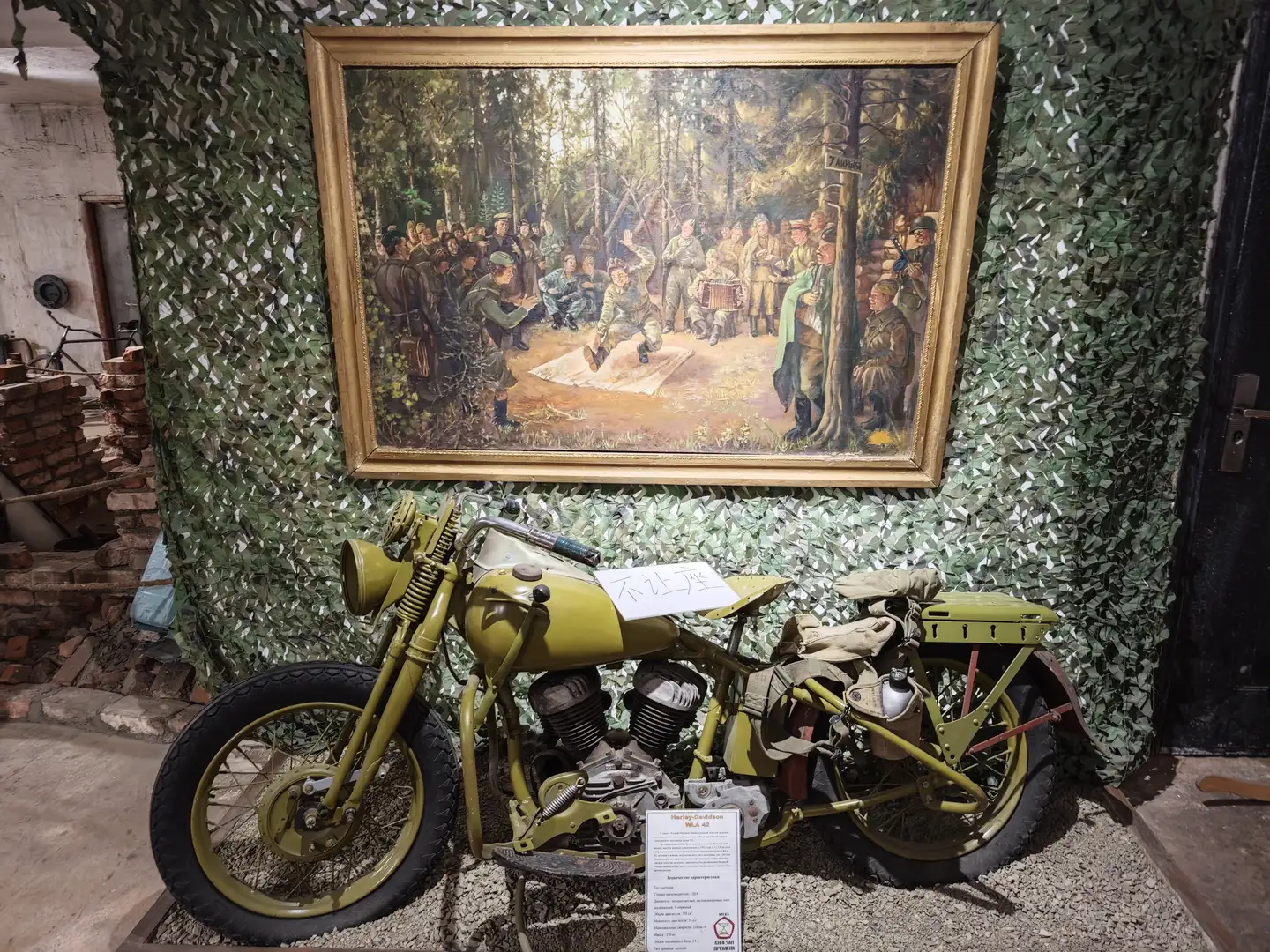 一幅描绘森林中人群聚集场景的油画挂在迷彩布背景墙上，下方停放着一辆黄色的老式军用摩托车，车身上贴有说明牌。