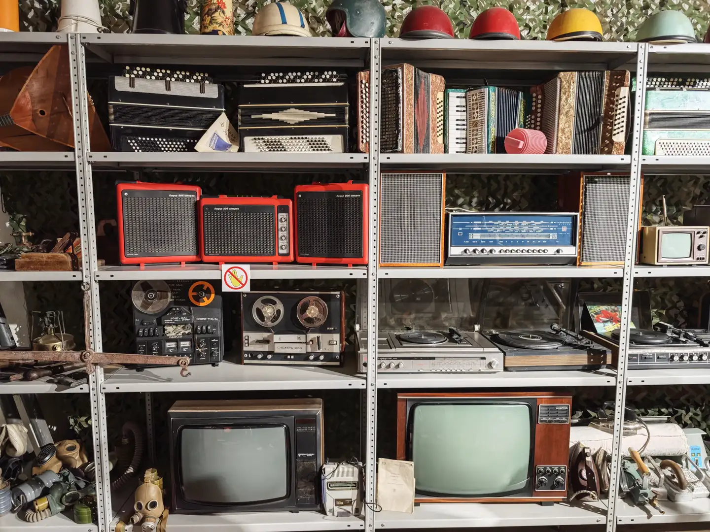 金属货架上整齐排列着各种复古电器，包括手风琴、收音机、录音机、唱片机和老式电视机，上方还摆放着不同颜色的头盔。