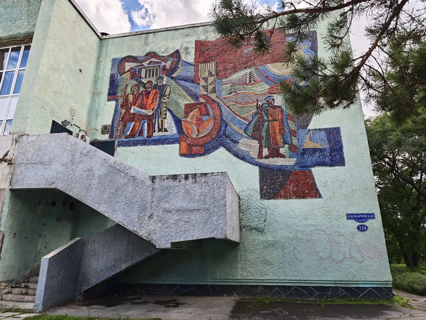 建筑物外墙上镶嵌着一幅大型马赛克壁画，描绘了人物和象征性图案，包括红旗、镰刀锤子和士兵形象，旁边有楼梯和树木。