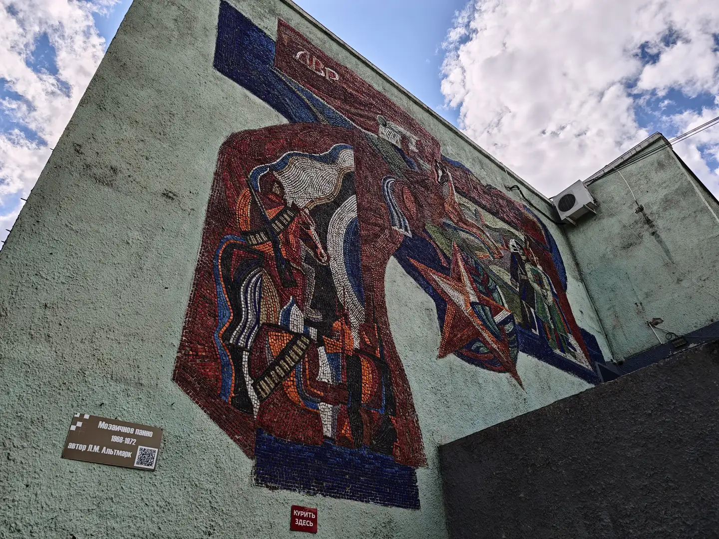 建筑外墙上有一幅大型马赛克壁画，画面中有骑马的战士、红星和旗帜，左下角标有“Мозаичное панно 1968-1972 автор Л.М. Альтмарк”字样，右侧有一个红色标志写着“КУРИТЬ ЗДЕСЬ”。