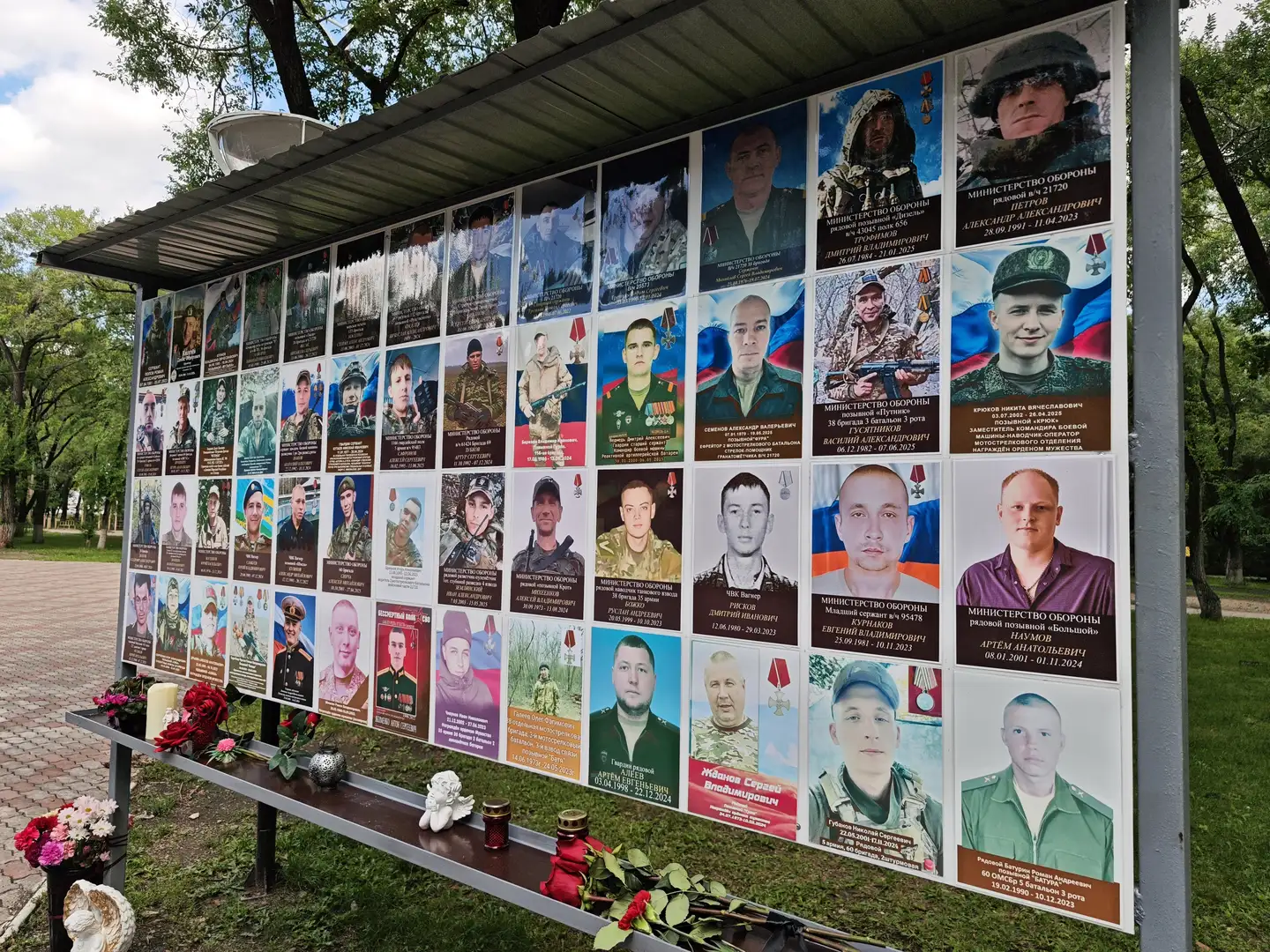 户外纪念墙上展示了许多军人的照片，每张照片下方有姓名和信息，墙前摆放着鲜花和蜡烛，周围是绿树和小路。