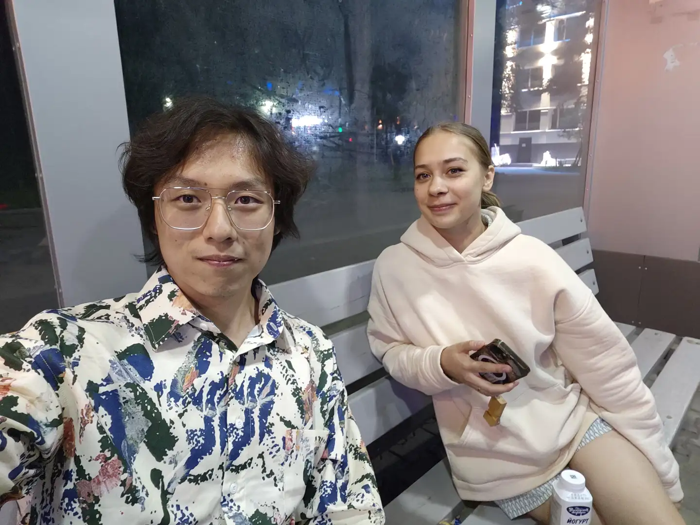两个人坐在长椅上，左边的人穿着花衬衫，右边的人穿着浅粉色连帽衫，手里拿着手机，旁边放着一瓶酸奶，背景是夜晚的玻璃窗。