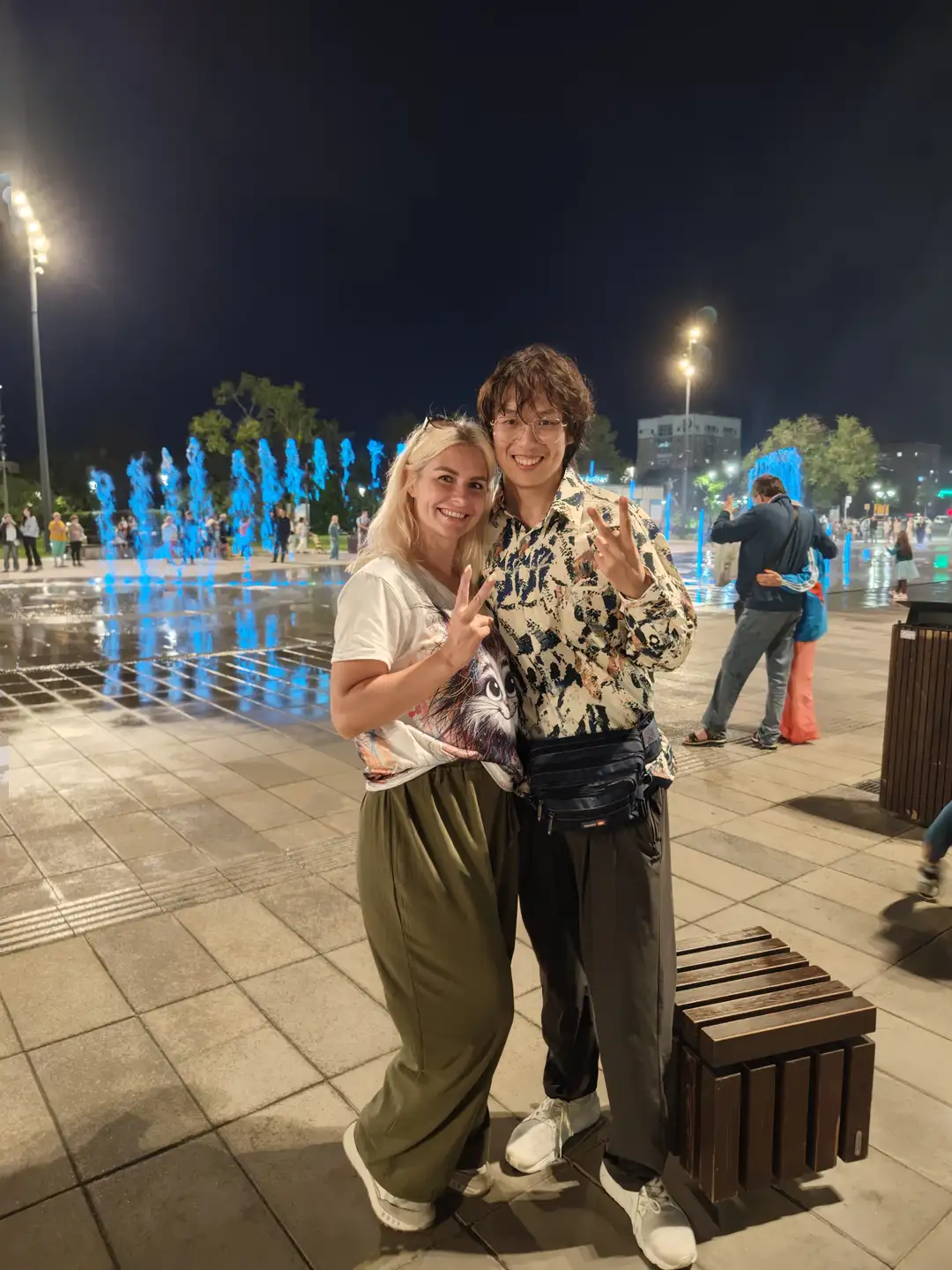两个人在夜晚的广场上合影，背景是蓝色灯光喷泉和一些行人。左边的人穿着白色T恤和绿色宽松裤子，右边的人穿着图案衬衫和黑色裤子，腰间挎着一个包。两人都面带微笑，比出胜利手势。