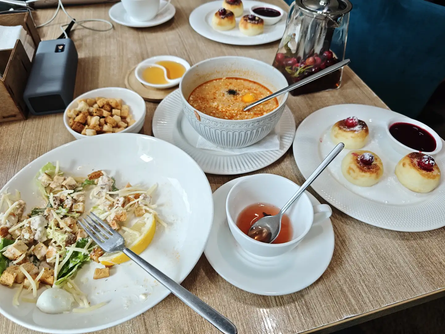 桌上摆放着凯撒沙拉、俄式罗宋汤、小点心和茶，旁边有面包丁和果酱，餐具整齐地放在盘子上。