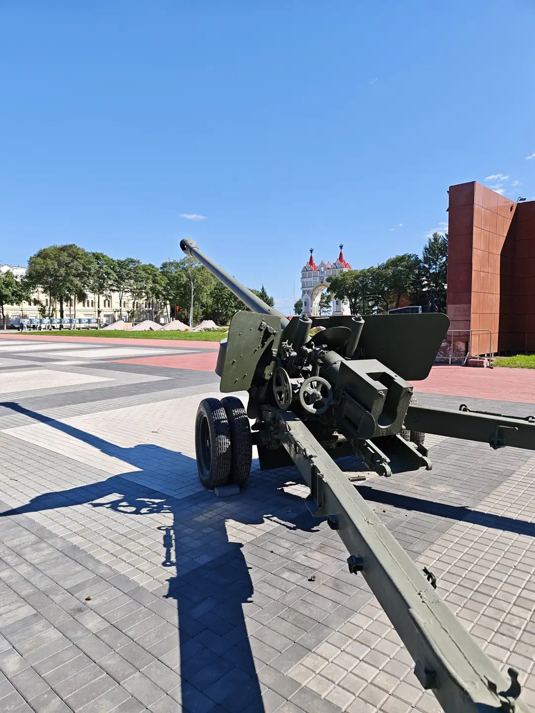 广场上展示着一门绿色的旧式火炮，背景是蓝天、树木和一座白色建筑，建筑顶部有红色装饰。