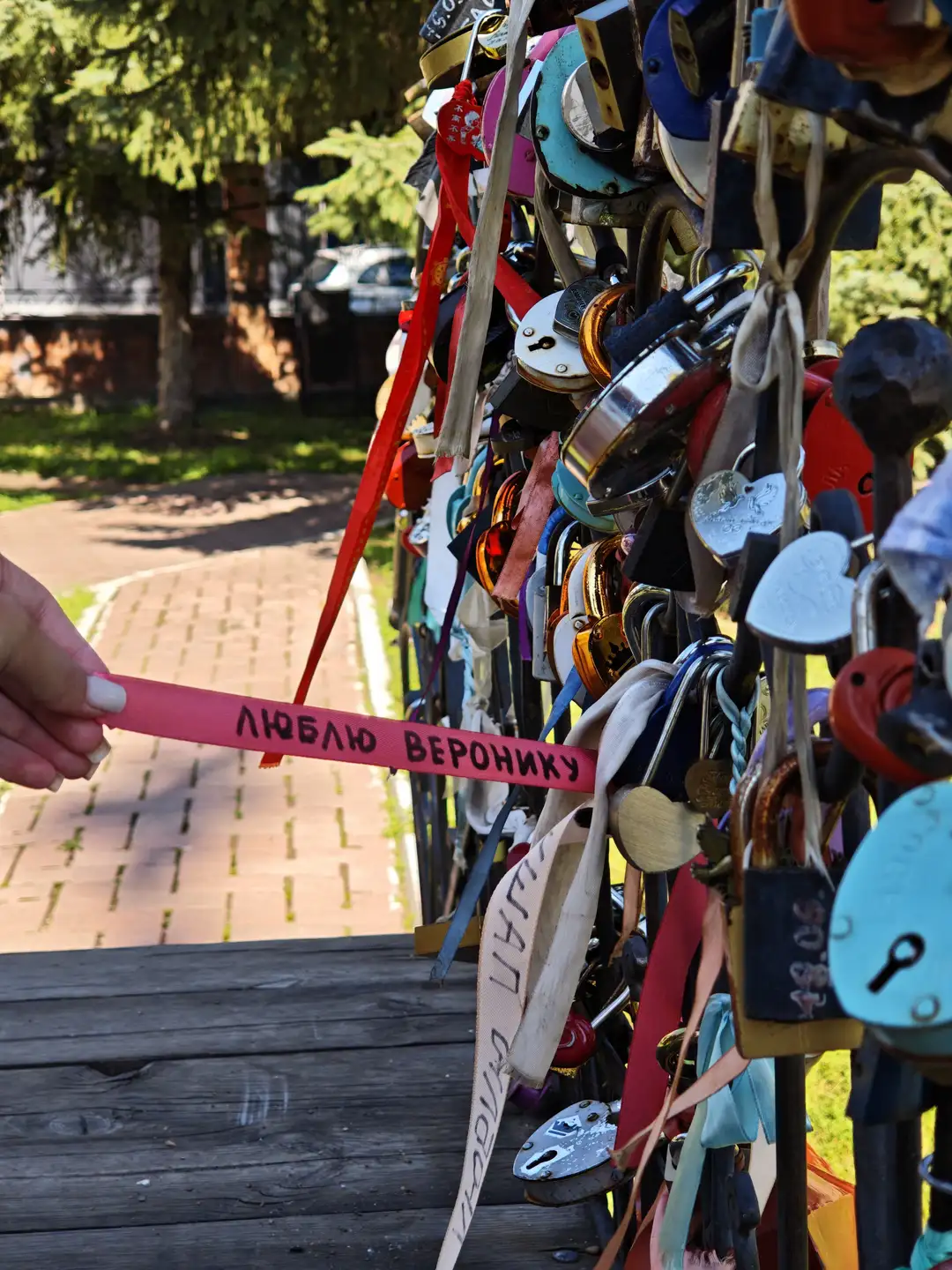 一只手拿着一条写有“люблю Веронику”的红色丝带，挂在布满彩色挂锁和丝带的栏杆上。