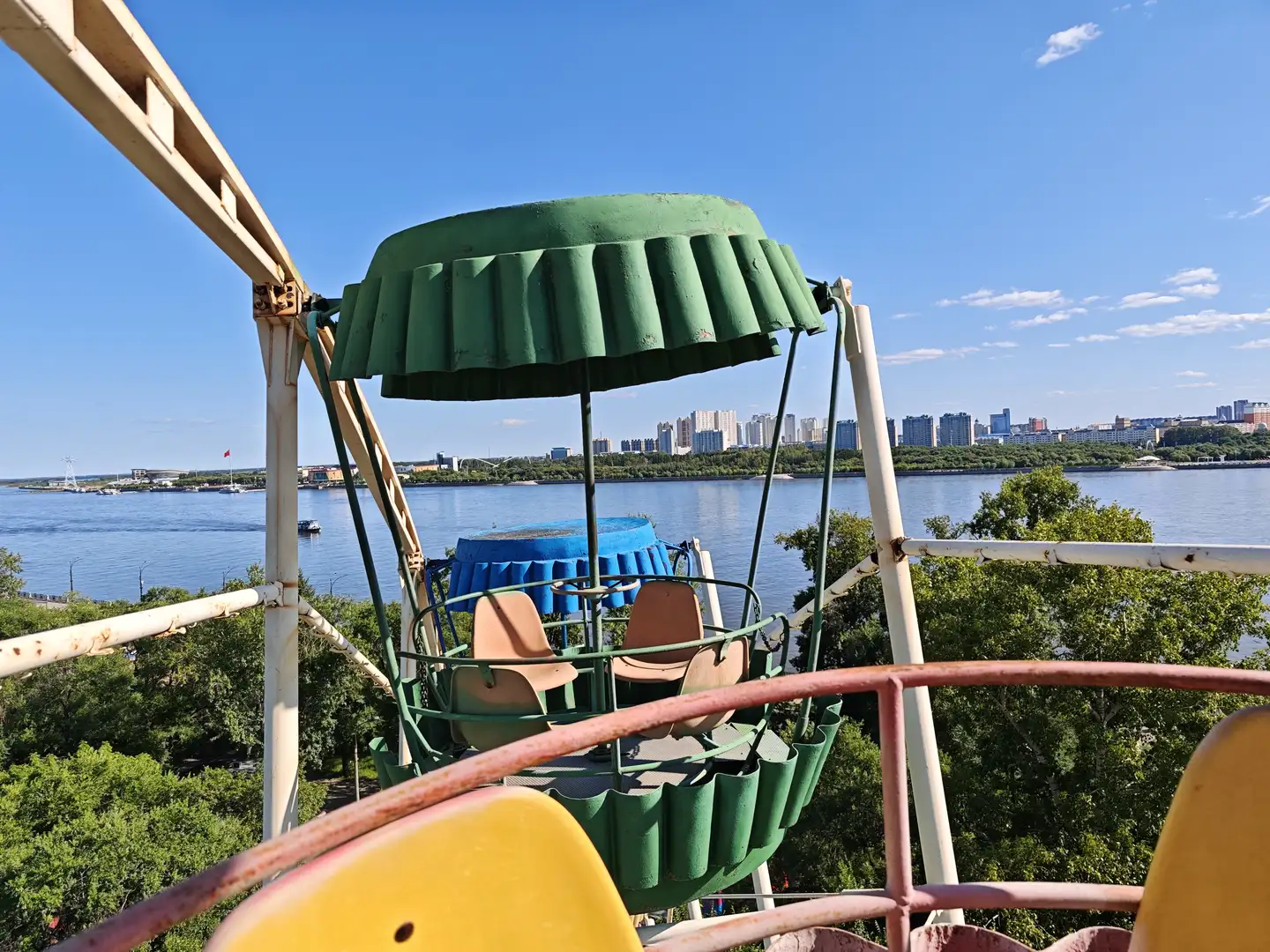 摩天轮的绿色座舱悬挂在高处，下方是蓝色的水面和远处的城市建筑群，天空晴朗。