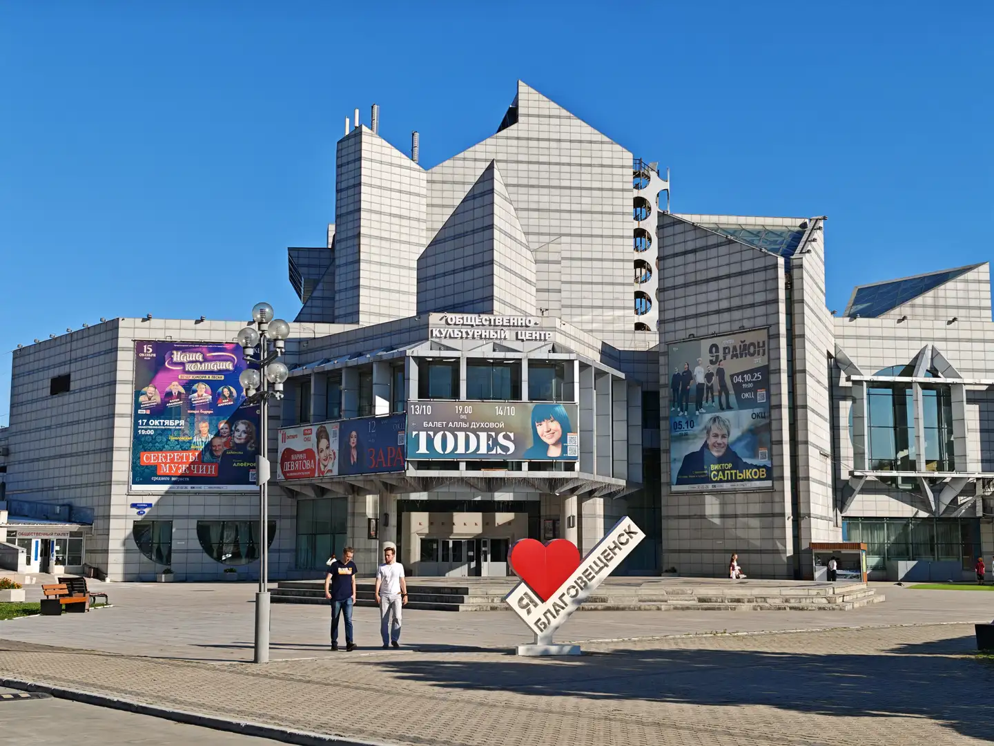 现代风格的文化中心大楼前立着一个红色心形标志，上面写着“Я ♥ Благовещенск”，大楼外墙上挂着多张演出海报。