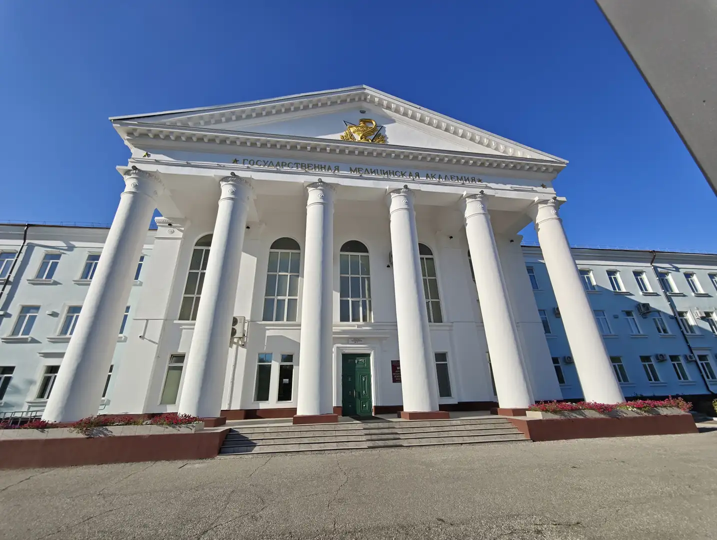 白色建筑正面有六根高大的圆柱，顶部刻有“ГОСУДАРСТВЕННАЯ МЕДИЦИНСКАЯ АКАДЕМИЯ”，门前有台阶和花坛。