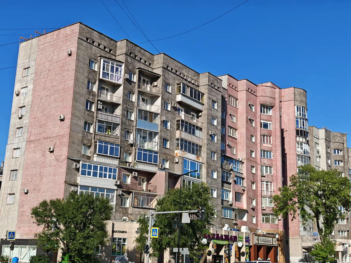 一栋多层住宅楼，外立面由粉色和灰色砖块构成，窗户排列整齐，部分阳台装有玻璃窗或金属栏杆。楼体底部设有商铺，招牌上有“ОCTABKA”等字样，门前有交通信号灯和行人过街标志。周围有绿树和电线，天空晴朗无云。