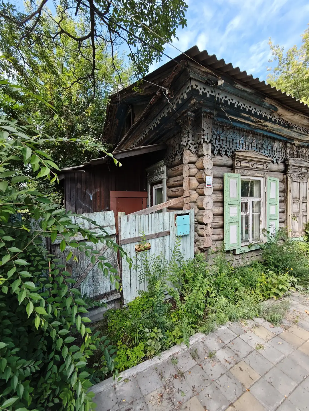 木制小屋由原木搭建，屋顶为波纹铁皮，窗户带有绿色百叶窗，门前有低矮的木栅栏和茂密的绿植。房屋部分结构陈旧，墙角有雕刻装饰，周围树木环绕，环境宁静。