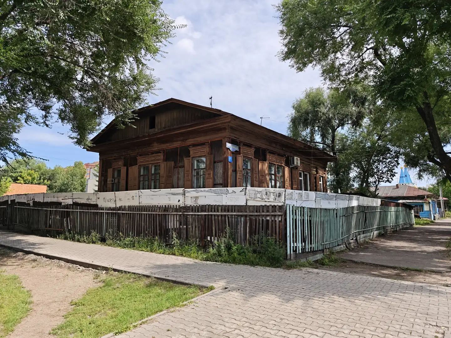 两层木质建筑位于街道旁，外立面为深棕色，多扇窗户整齐排列，前方有白色与蓝色相间的围栏。建筑后方可见树木和远处的尖顶建筑，天空晴朗，云朵稀疏。
