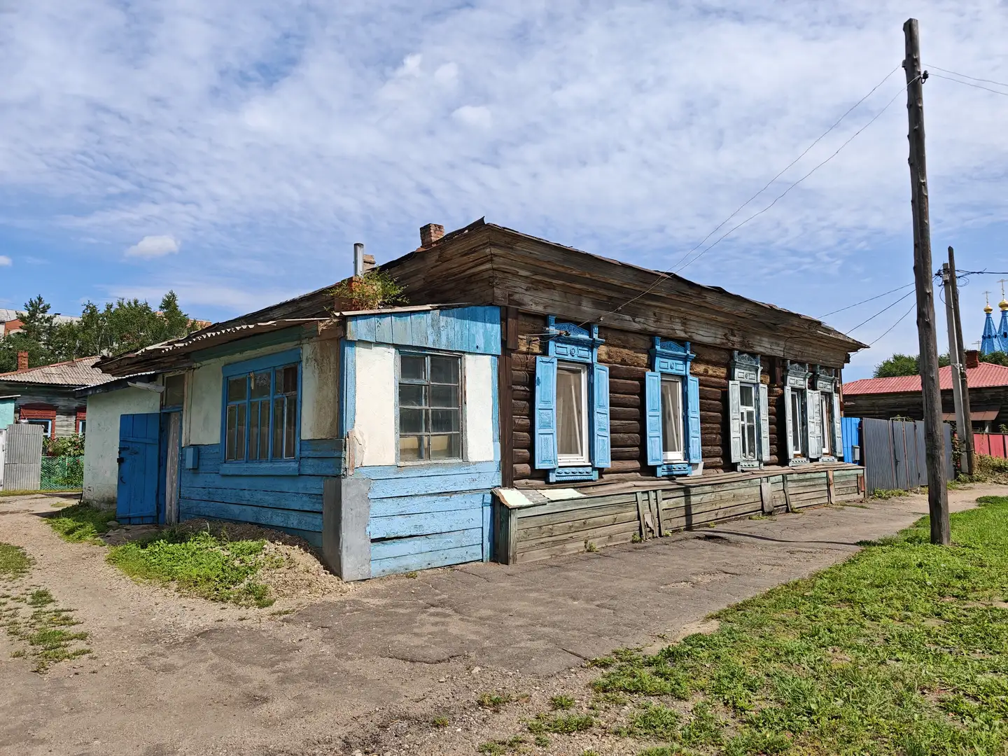 一栋蓝白相间的木结构房屋，墙体部分为原木，部分涂成蓝色，窗户装有蓝色窗框。房屋前有一条水泥路，旁边是草地，背景中可见电线杆和远处的红色屋顶建筑。