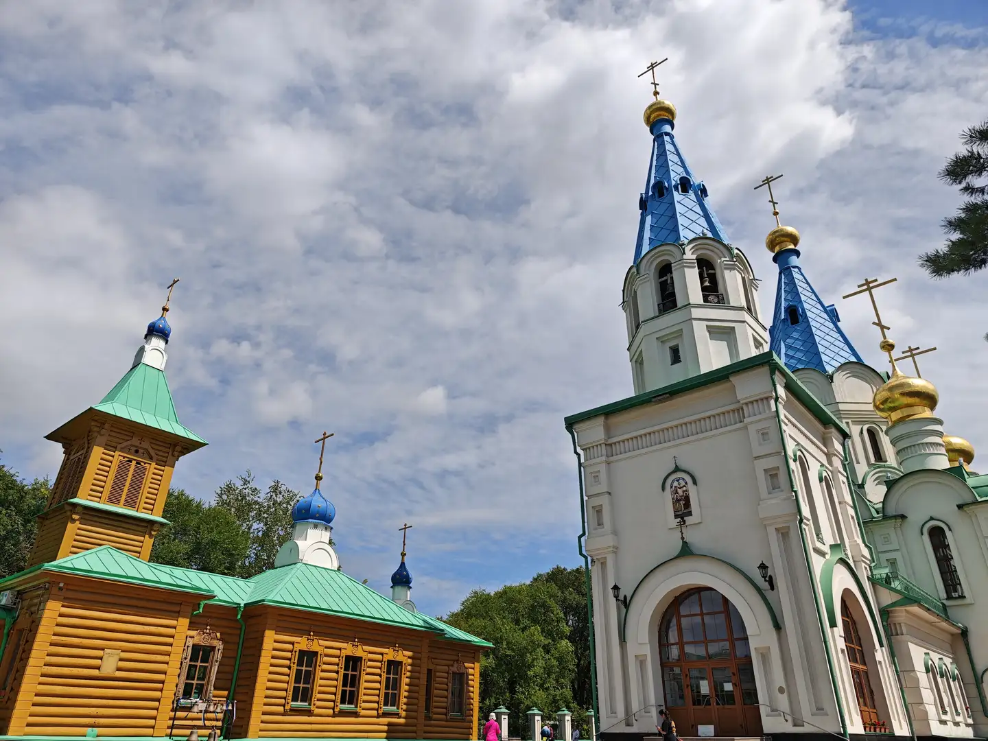 一座白色教堂建筑，顶部有蓝色尖顶和金色圆顶，十字架高耸，旁边是一座黄色木制建筑，屋顶为绿色。天空中有白云，建筑周围有树木，前景有人在行走。