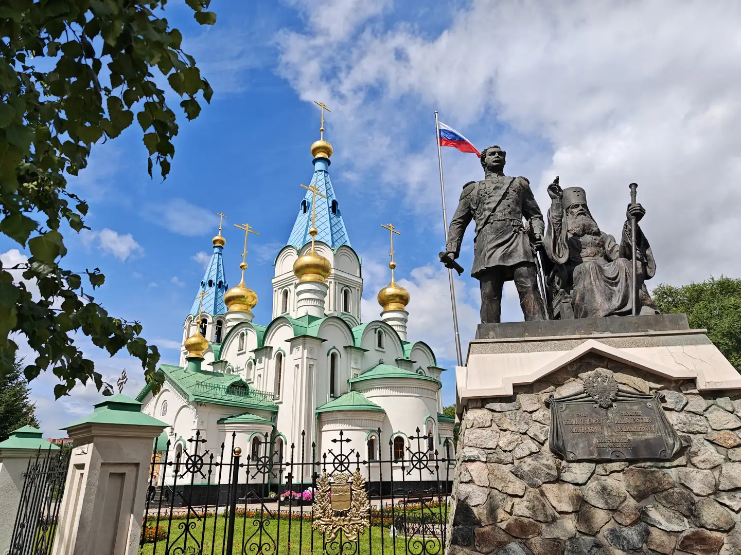 一座大型白色教堂，拥有多个蓝色和金色圆顶，前方矗立着一座雕像，雕像上有两名人物，一人站立持剑，一人坐着持权杖。教堂前有黑色铁艺围栏，俄罗斯国旗在风中飘扬。