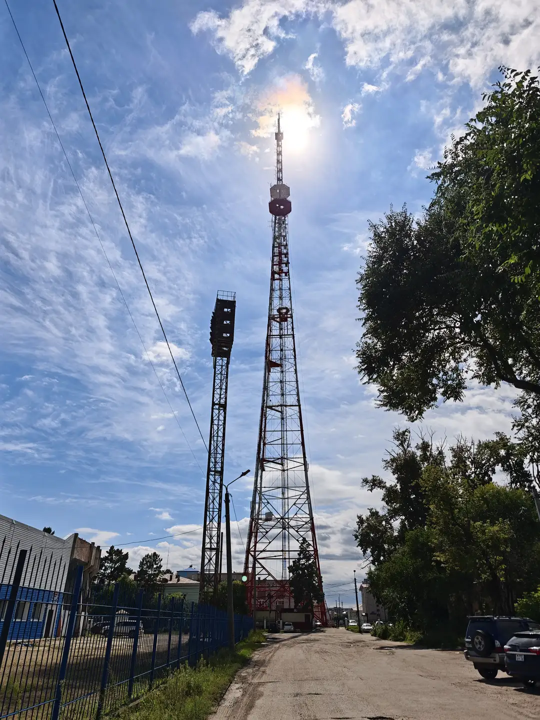 一座高耸的红白相间通讯塔，塔身呈三角形结构，顶部有信号设备。塔旁是一条小路，路边有树木和围栏，远处可见建筑物和停放的车辆，阳光从塔顶上方照射下来。