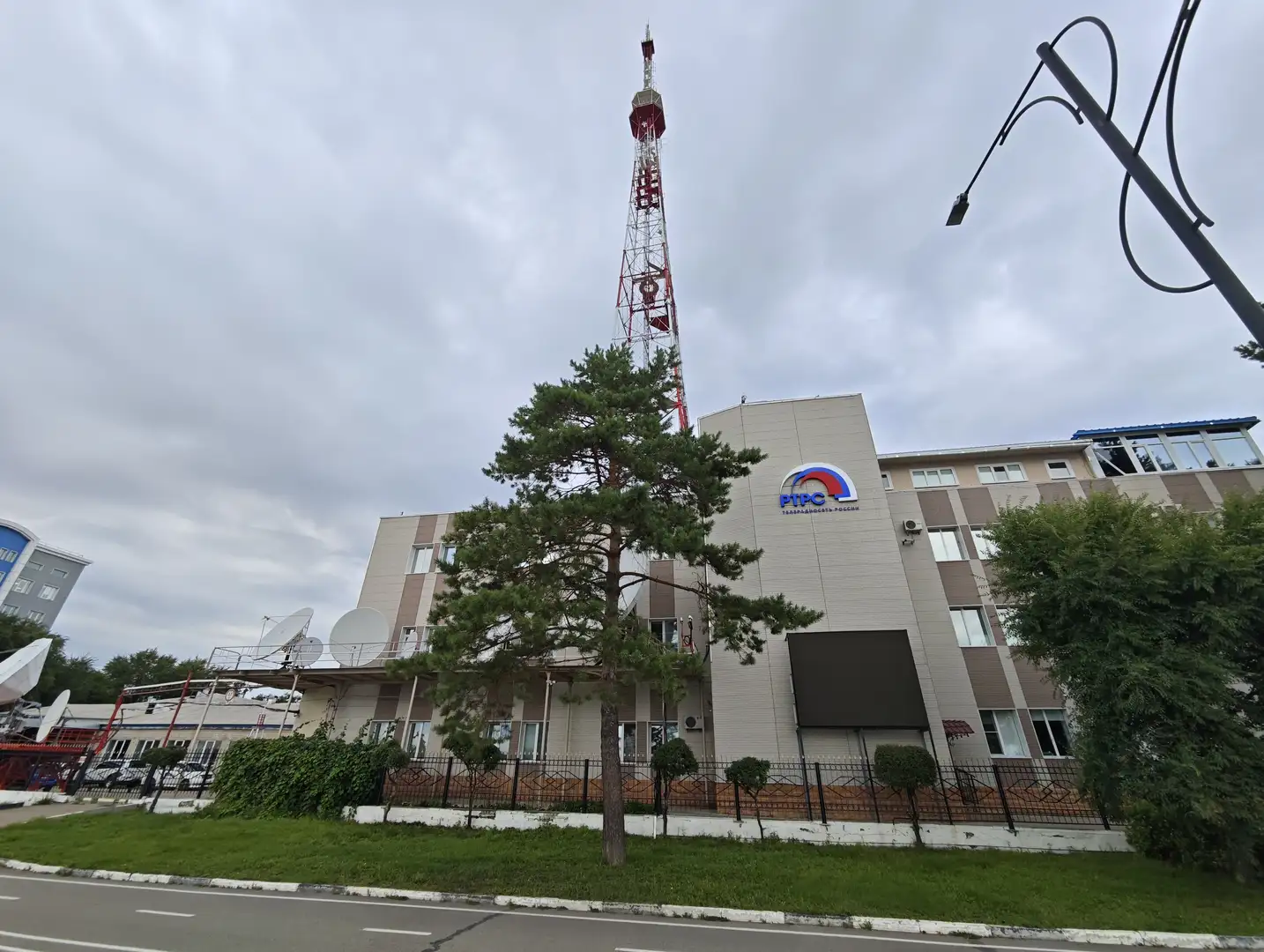 一栋现代建筑，正面有“РТРС”标志，建筑前有树木和草坪，旁边有一座高大的红白相间通讯塔。建筑屋顶安装了多个卫星天线，周围有围栏，天空阴沉。
