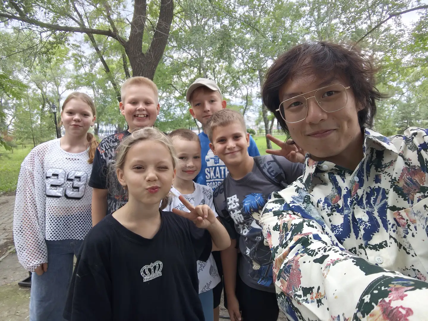 一群孩子和一名戴眼镜的成年人在户外合影，背景是树木和公园小径。孩子们穿着休闲服装，有的比出胜利手势，表情自然。成年人身穿花纹衬衫，站在最右侧。