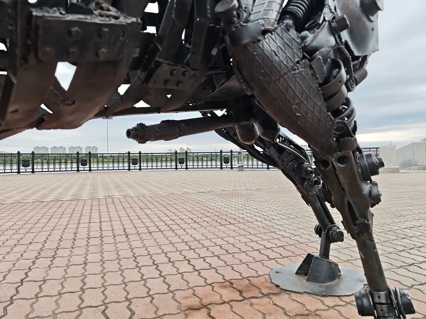 一个由金属部件构成的机械结构，底部有类似腿的支撑，安装在铺有砖块的地面上，背景是栏杆和远处的建筑群