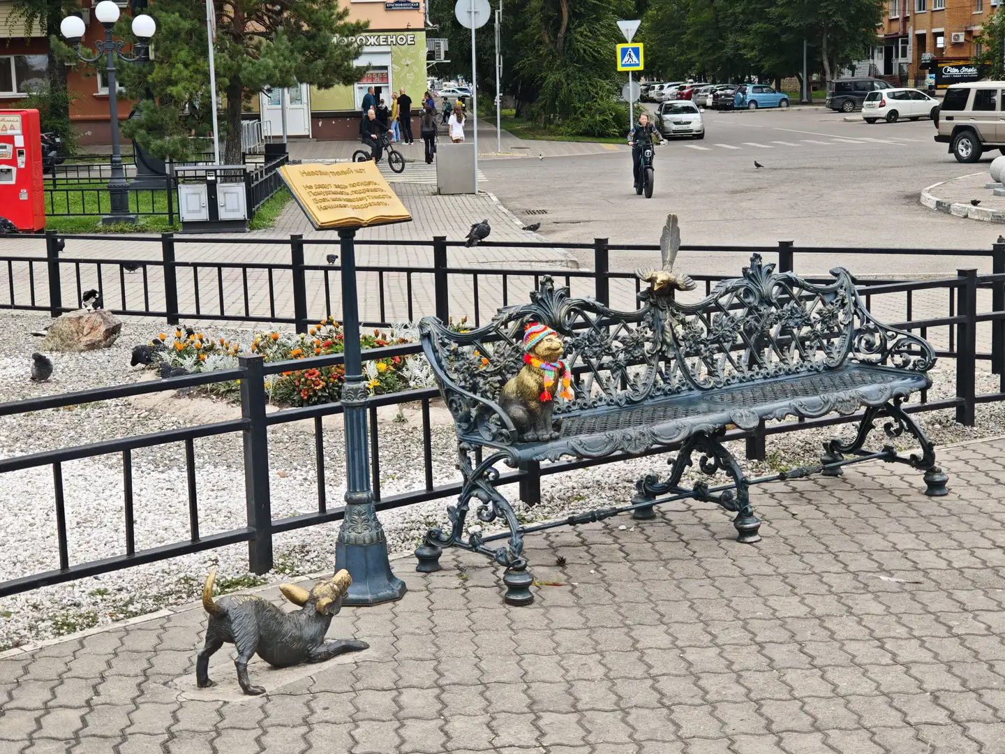 一张装饰性长椅，上面有一只戴着彩色围巾的猫雕像，旁边是一只狗雕像，周围有花坛、栏杆和街道，背景中有人行走和骑车