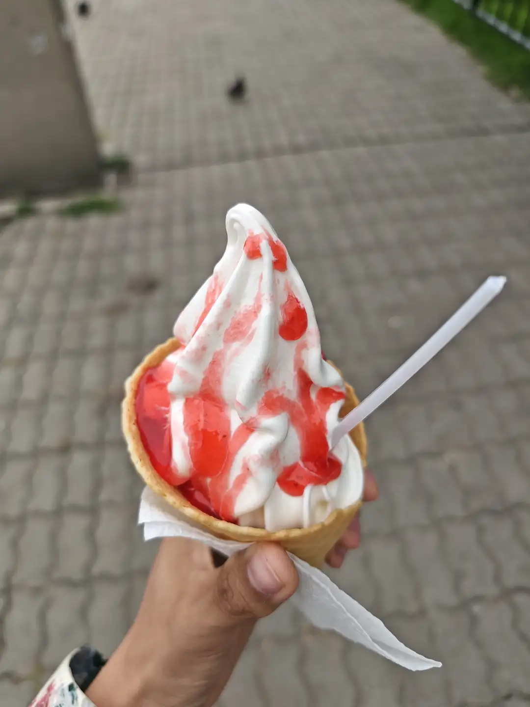 一只手拿着一个冰淇淋蛋筒，里面装着白色软冰淇淋，上面淋有红色果酱，蛋筒用纸包裹，插着一根塑料勺子