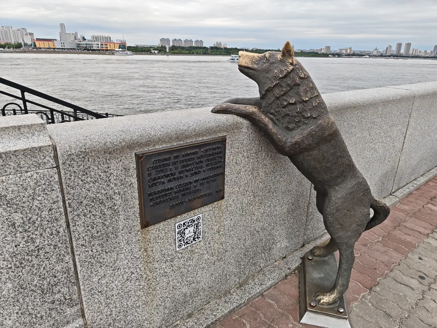 一只青铜狗雕像靠在石墙上，前爪搭在墙沿上，面向水面，墙上有一块铭牌和一个二维码，远处是城市天际线和河流