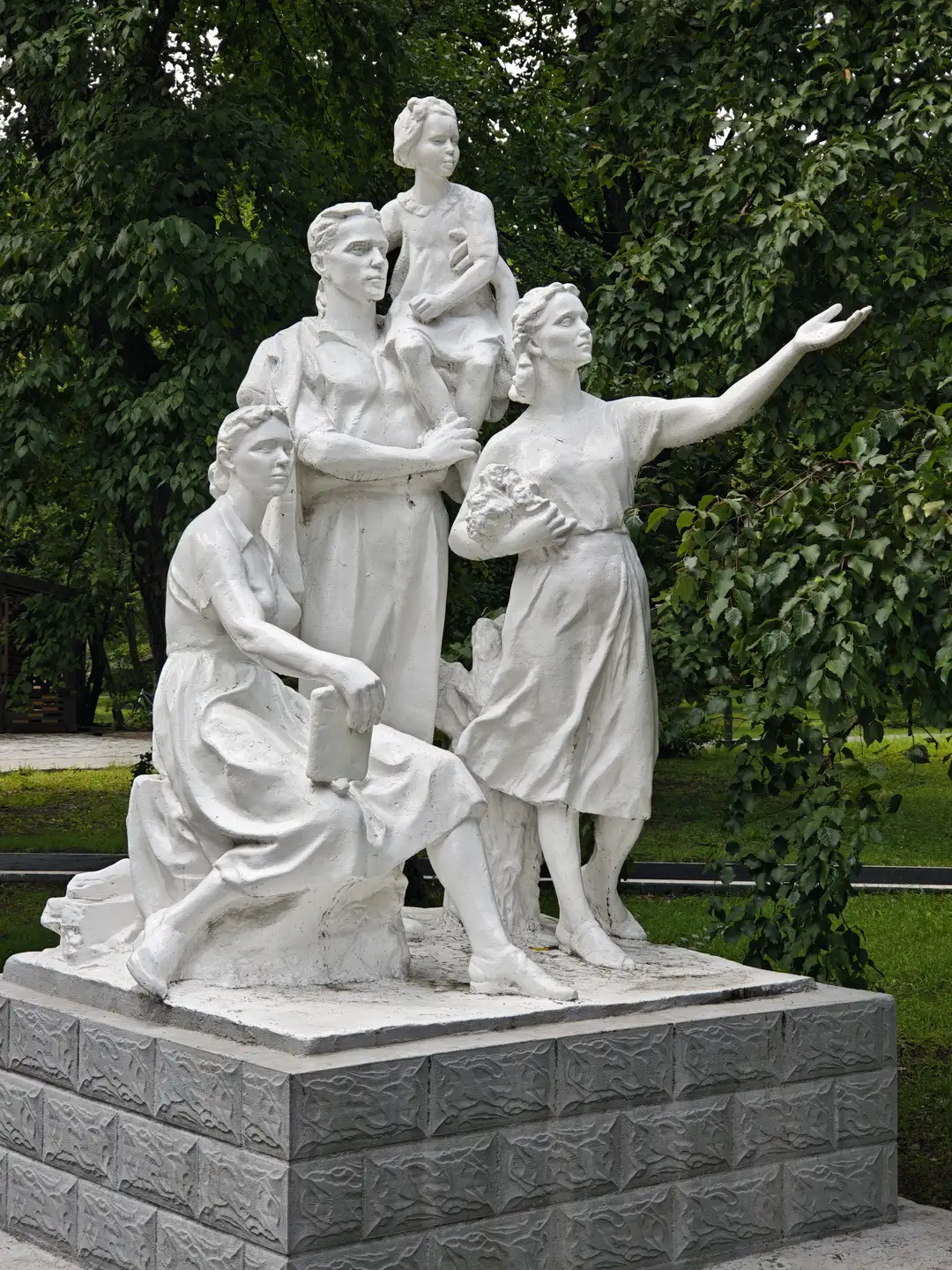 一座白色石雕展示四个人物，其中一人跪坐持书，另一人站立伸出手臂，第三人抱着孩子，第四人站在中间。雕塑位于户外，周围有绿树环绕，底座为方形石基。