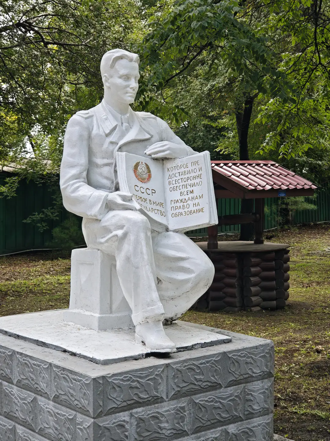 一座白色雕像描绘一个穿着制服的男子坐在石墩上，手中捧着一本打开的书，书上有俄文文字和苏联国徽。雕像置于公园环境中，背景有树木、木制凉亭和绿色围栏。