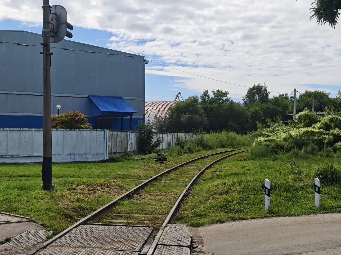 两条铁轨从前景延伸至远处，穿过一片草地，旁边是一栋灰色工业建筑和蓝色遮阳棚。轨道旁有白色护栏和交通信号灯，天空多云，远处有树木和电线杆。