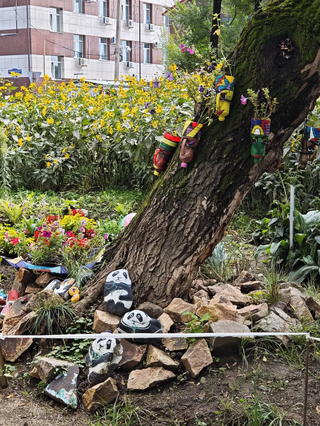 一棵大树旁的花坛中种满了五颜六色的花朵，树干上挂着多个彩绘塑料瓶，瓶中种有植物。树根周围堆着石头，上面画有熊猫图案，背景是居民楼和绿植。