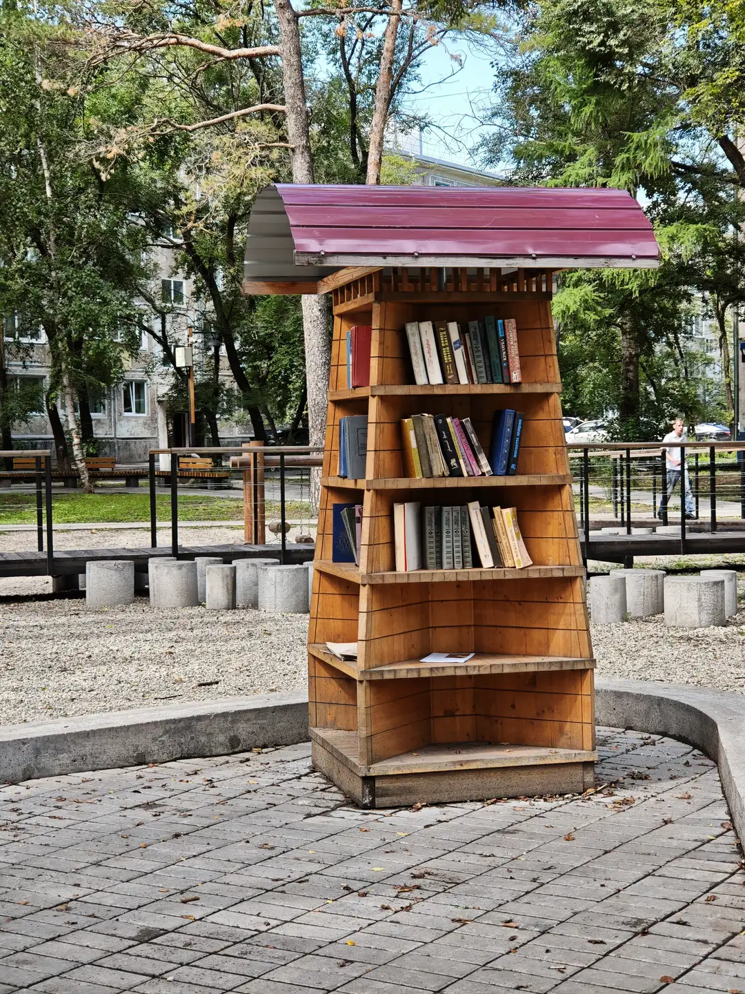 一个木制书架立在铺有石砖的地面上，顶部有红色金属屋顶，书架内摆放着多本书籍。周围是树木、长椅和行人，背景有建筑物和围栏。