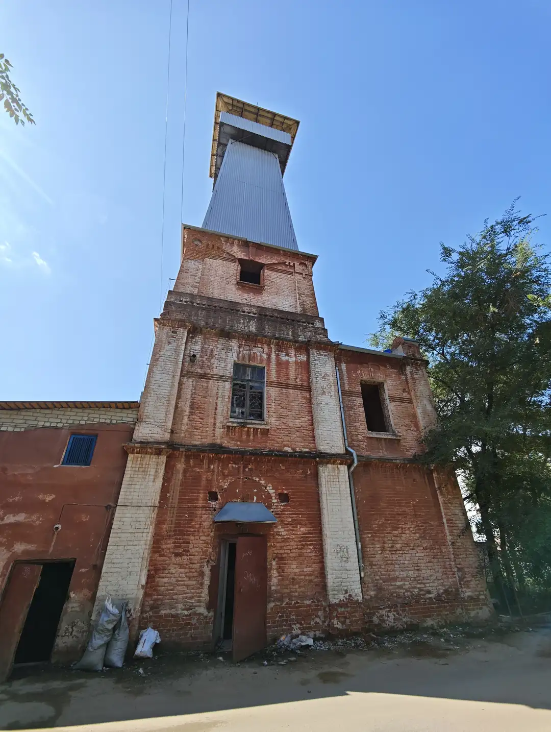 一座红砖砌成的高塔建筑，顶部有金属结构和方形开口，墙体斑驳，部分窗户破损。塔旁有树木和杂物堆积，地面散落垃圾，显得有些荒废。