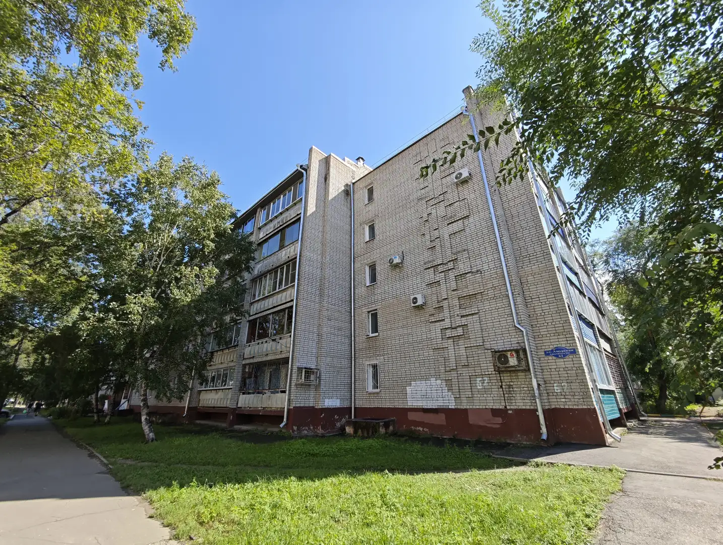 一栋多层住宅楼，外立面为浅色瓷砖，部分区域有剥落痕迹。楼体周围绿树成荫，底层有空调外机和红色基座，旁边是人行道和草地。