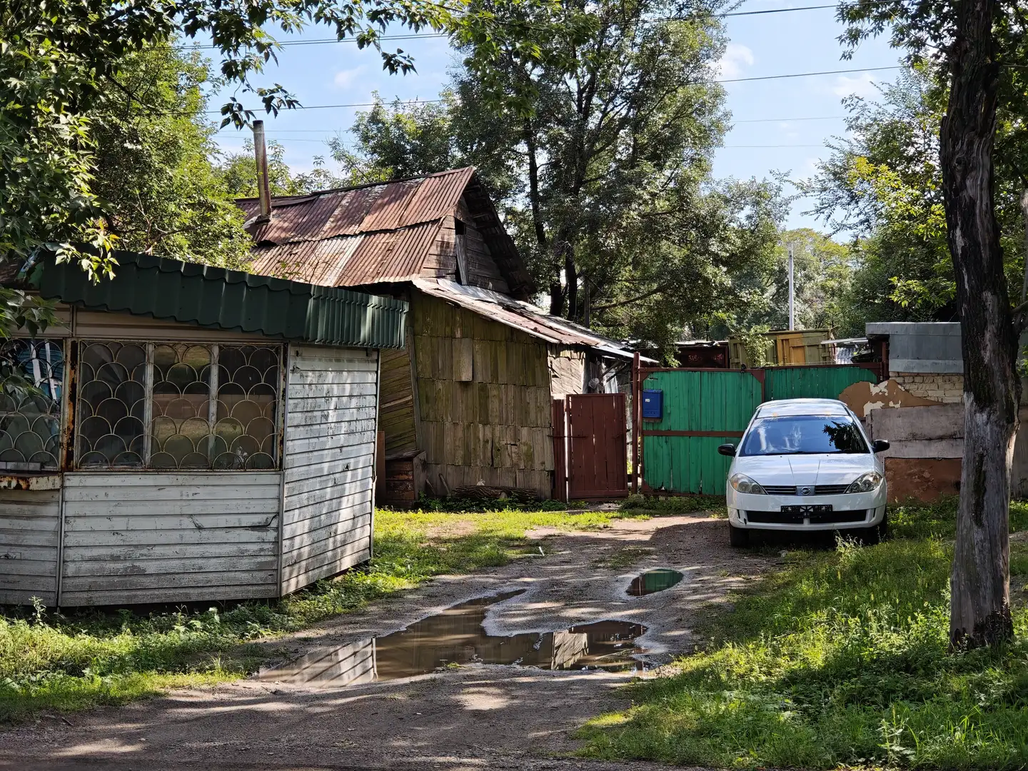 一条泥泞的小路穿过几间老旧木屋，屋顶锈迹斑斑，墙面陈旧。路边停着一辆白色轿车，周围绿树环绕，地面有积水反光。
