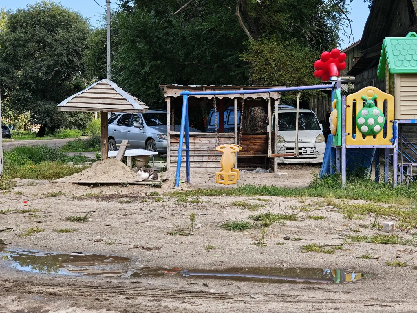 一个儿童游乐场，设有秋千、滑梯和沙坑，地面为沙土，有水坑。远处停着几辆车，周围有树木和简易棚屋，一只猫卧在沙堆旁。