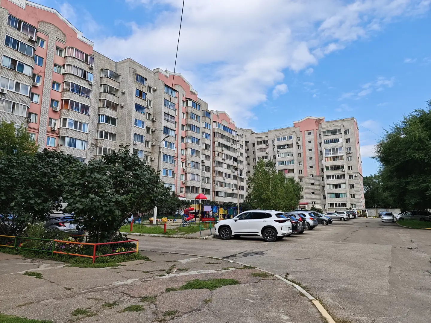 一排高层住宅楼，外立面为灰白与粉红相间，楼前有停车场和绿化带。天空晴朗，云朵稀疏，楼间可见儿童游乐设施和停放的车辆。