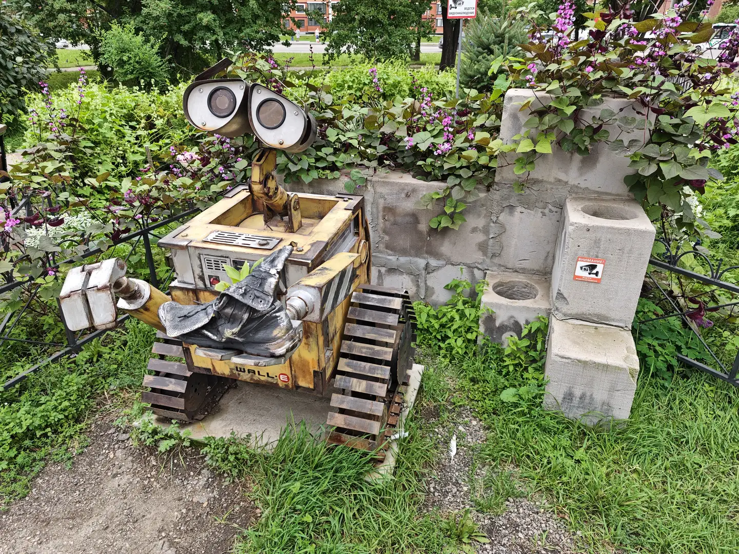 一个用金属和木材制作的机器人雕塑，外形类似动画角色WALL·E，放置在花坛中。周围有绿色植物和紫色花朵，旁边是水泥块堆砌的矮墙。