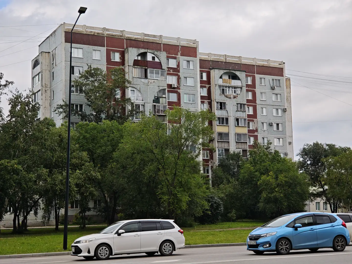 一栋多层住宅楼，外立面为灰色和红色相间，部分墙面剥落。楼前有树木和草坪，道路上行驶着白色和蓝色的汽车，天空阴沉。