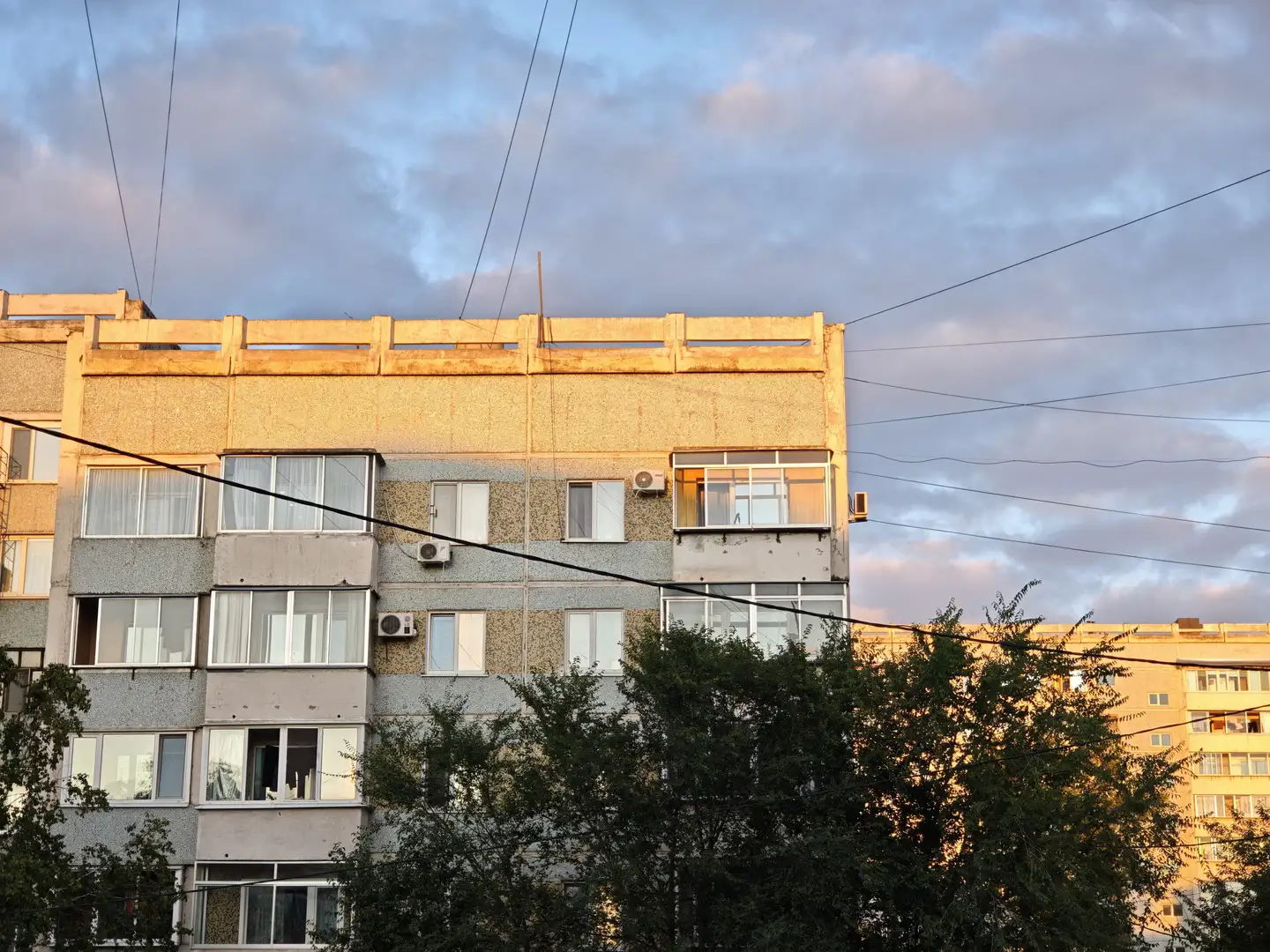 一栋多层住宅楼的顶部在夕阳照射下泛着金黄色光芒，窗户排列整齐，部分装有空调外机。建筑外墙为浅色与深色相间，屋顶边缘有护栏结构。天空中布满灰蓝色云朵，电线横跨画面。