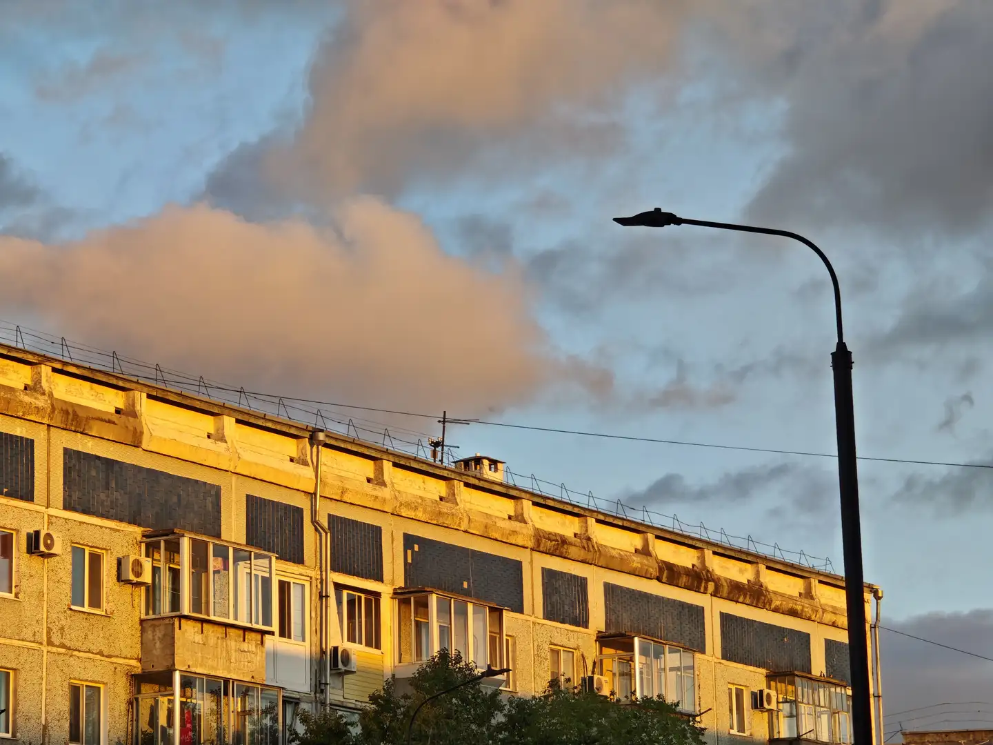 一排老旧住宅楼的侧立面被夕阳染成暖黄色，屋顶上设有金属围栏和小型通风口。窗户多数为封闭式，部分带有玻璃阳台，外墙上安装多个空调外机。右侧有一根高耸的路灯杆，背景是蓝灰色的天空和蓬松的云层。