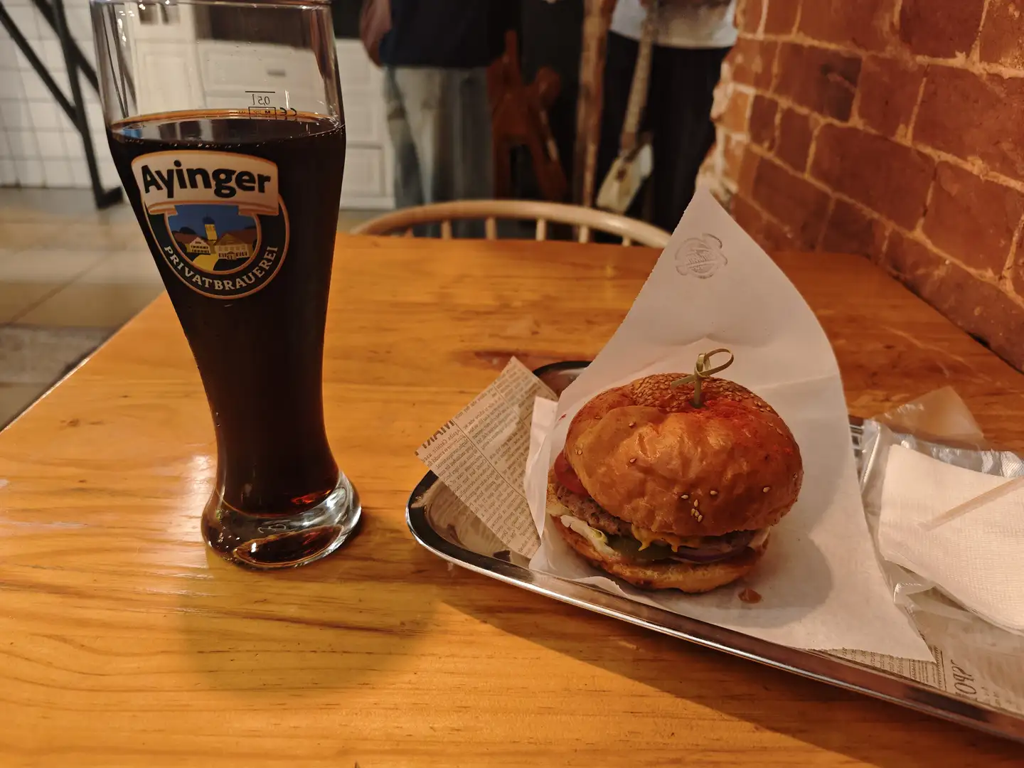 木桌上摆放着一杯深色啤酒和一个汉堡，啤酒杯上有“Ayinger PRIVATBRAUEREI”的标志。汉堡放在金属托盘上的纸巾中，夹有生菜、番茄和芝士，用牙签固定。背景可见砖墙和模糊的人影，环境像是餐厅或酒吧内部。