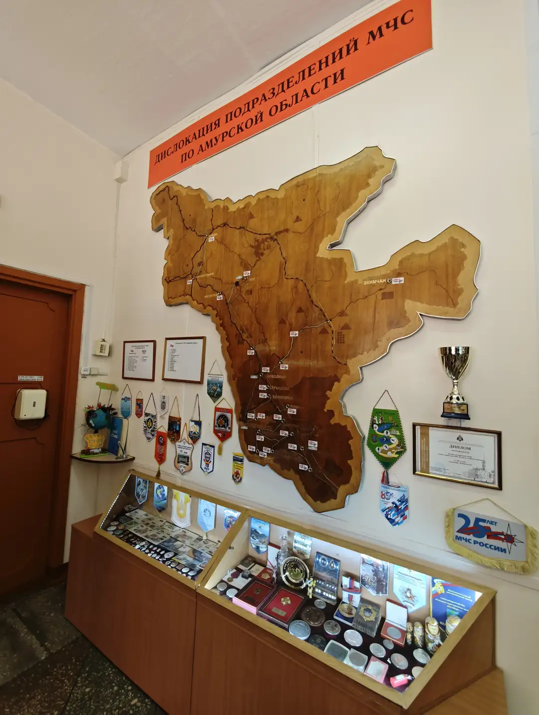 墙上挂着一幅大型木质地图，显示了阿穆尔州的应急管理部门分布情况。地图下方陈列着多个徽章和奖牌，旁边还有一座奖杯和证书。展柜中摆放着各种勋章、证书和纪念品，展示了该地区的应急管理历史。