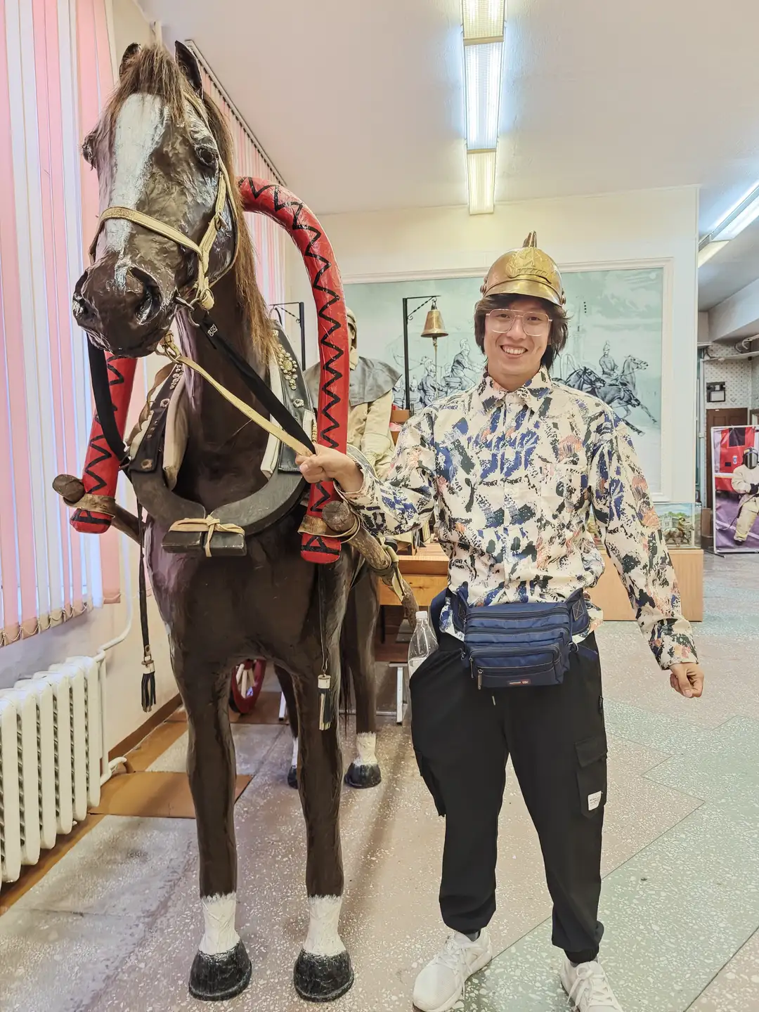 一位穿着花衬衫和黑色裤子的人站在一匹装饰华丽的马旁边，头戴金色头盔，面带微笑。马身披有红色装饰物，牵着缰绳，背景是粉白相间的墙壁和一幅描绘骑马场景的画作。房间内还有其他展品和展示架。