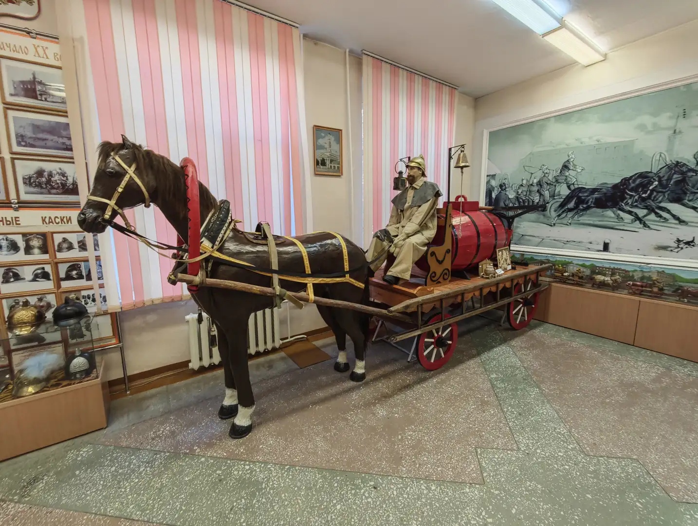 一匹棕色的马模型拉着一辆红色的消防车，车上坐着一个穿着旧式消防制服的人物模型。马匹佩戴着缰绳和装饰，车轮为红色，车厢上有一个大水桶。背景墙上有历史照片和一幅大型壁画，描绘了消防员骑马的场景。
