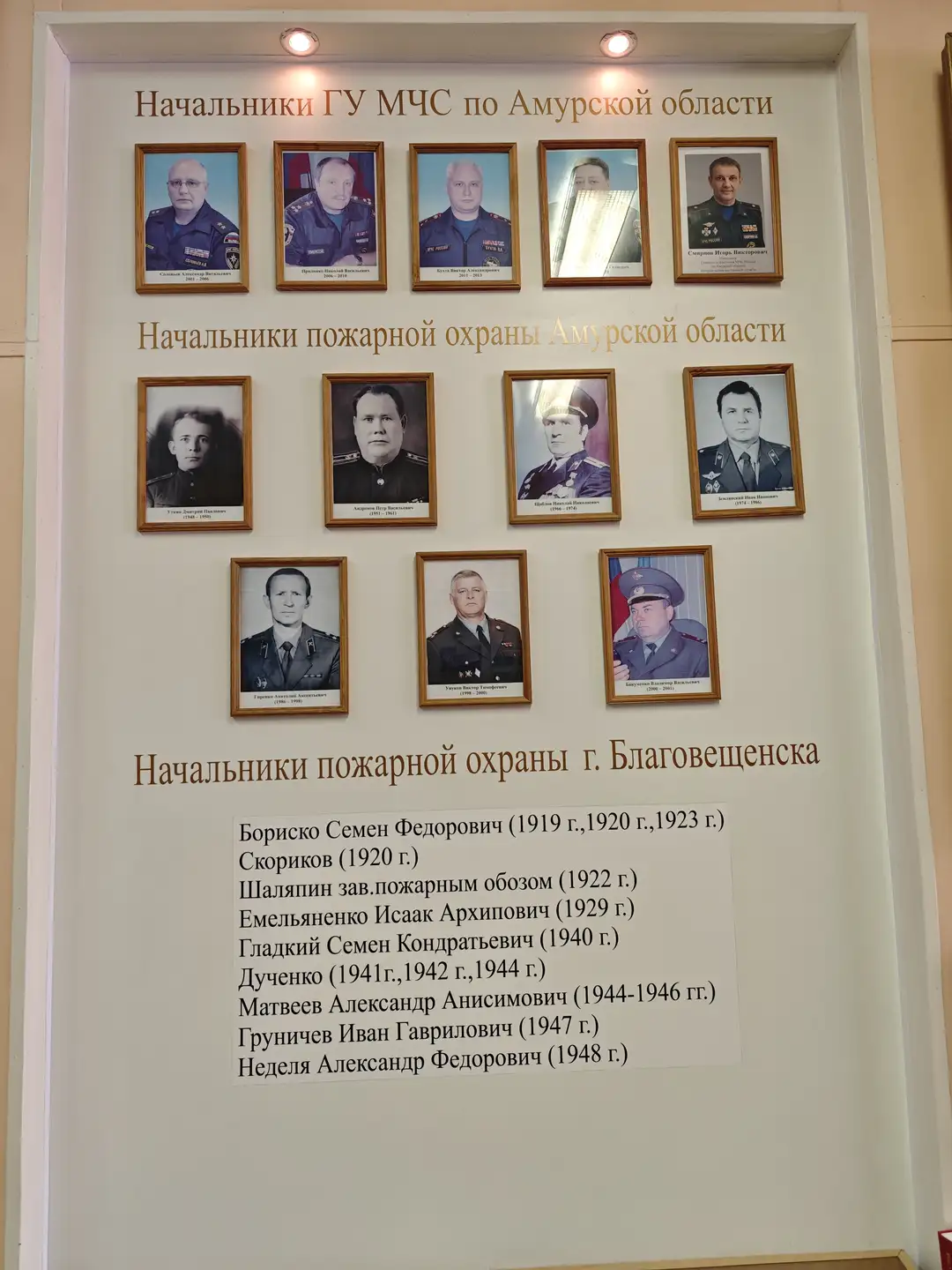 墙上展示着多张人物肖像照，分为三行排列，上方标注了“阿穆尔州紧急情况部负责人”等字样。每张照片下方有姓名和任职时间，最下方列出了一列文字，记录了布良斯克市消防部门历任负责人的名字和任期年份。整体布局整齐，灯光柔和。