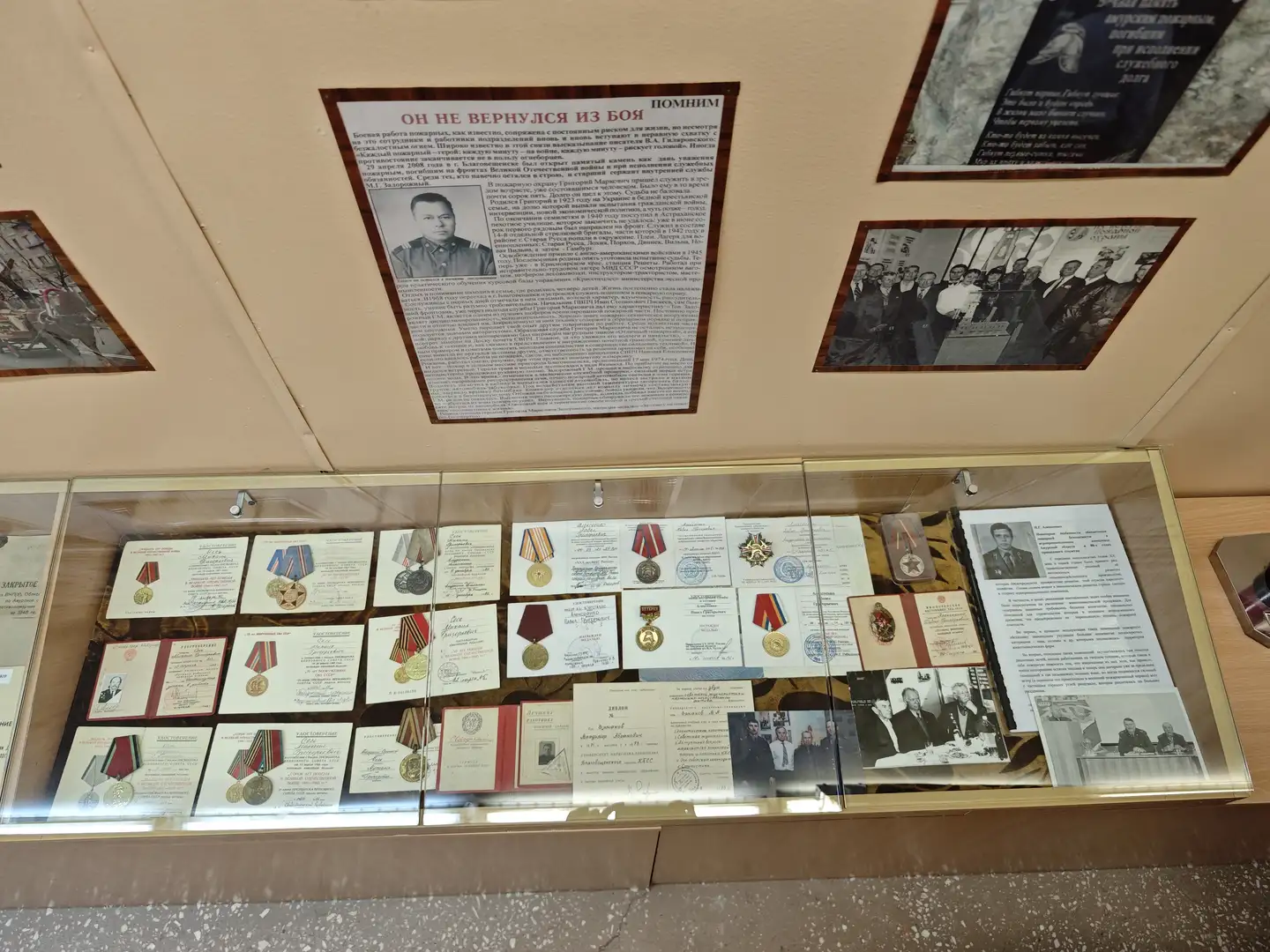 展柜中陈列着多枚勋章、奖章以及相关证书，每件物品都附有说明卡片。上方悬挂着一块写有俄文的展板，内容涉及某位未归来的英雄事迹。展柜内的物品按顺序排列，背景为浅色墙面，整体显得庄重而肃穆。
