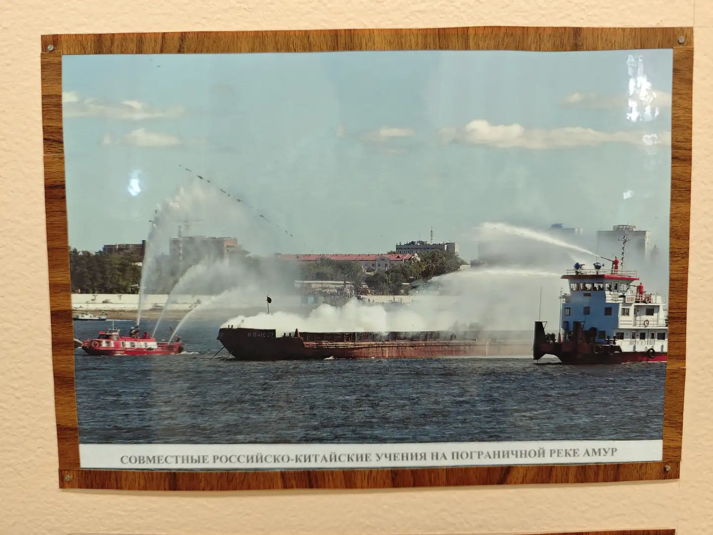 墙上挂着一幅装裱好的照片，展示的是中俄联合在阿穆尔河边境进行的消防演习。画面中有两艘船只正在向一艘货船喷水，背景是城市建筑和蓝天白云。照片下方有俄文标题，说明这是中俄联合演习的场景。